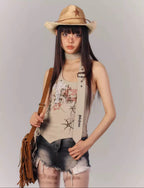 Woman wearing a beige sleeveless top with graphic design, denim shorts, and a straw hat on a gray background.